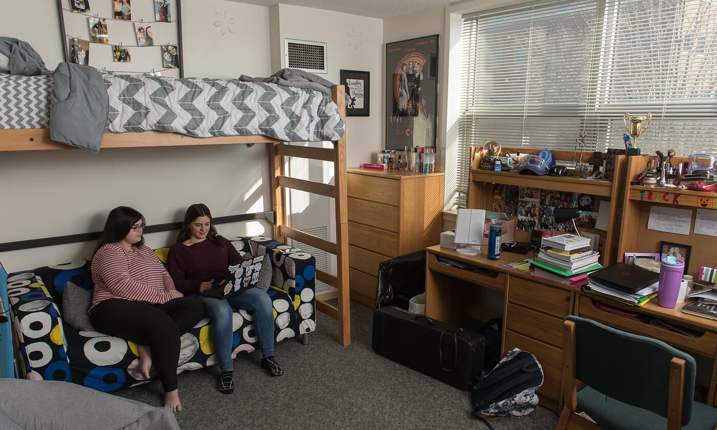 dorm room with two female students sitting at the bottom of a two-person bunk bed, looking at a laptop