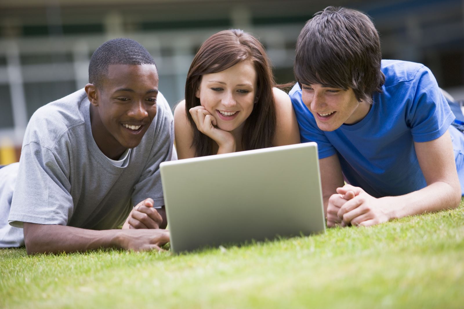 three students laying on the grass and watching a laptop screen